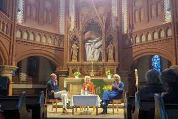 Zwei weiblich und eine männlich gelesene Person sitzen im Altarraum einer Kirche vor Publikum. 