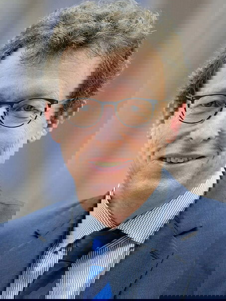 Das Bild zeigt eine männlich lesbare Person ohne Bart mit dunklen kurzen lockigen Haaren, einer Brille mit dunkler Fassung, einem hellblauen Hemd sowie einer dunkelblauen Krawatte und einem dunkelblauem Jackett.