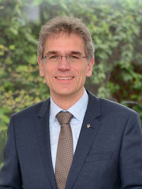 Eine männlich lesbare Person ohne Bart, mit Brille sowie kurzen grauen Haaren, mit hellblauem Hemd, brauner Krawatte und dunkelblauem Sakko.