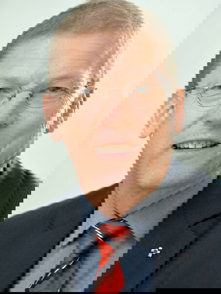 Eine männlich lesbare Person ohne Bart, mit Brille und kurzen Haaren, in mittelblauem Hemd und dunkelblauem Sakko sowie mit einer roten Krawatte.