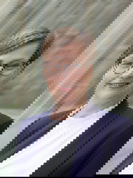 Eine weiblich lesbare Person mit kurzem braunen Haar, einer braunen Brille, weißem Oberteil und dunkelblauem Jackett.