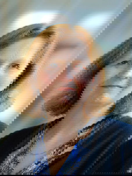 Eine weiblich lesbare Person mit schulterlangem rotblondem Haar, einem blau-gemusterten Oberteil und einer dunkel blauen Jacke.