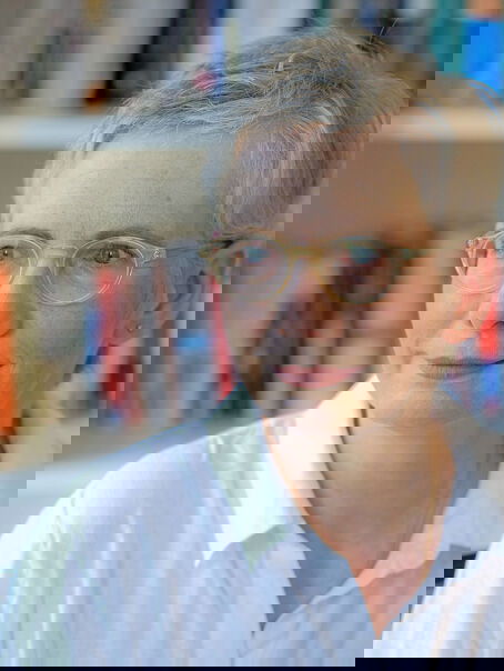Der Bild zeigt eine weiblich lesbare Person mit kurzen hellblonden Haaren, einer hellen Brille, heller Hautfarbe und einer weißen Bluse. Die Person sitzt vor einem gefüllten Bücherregal.
