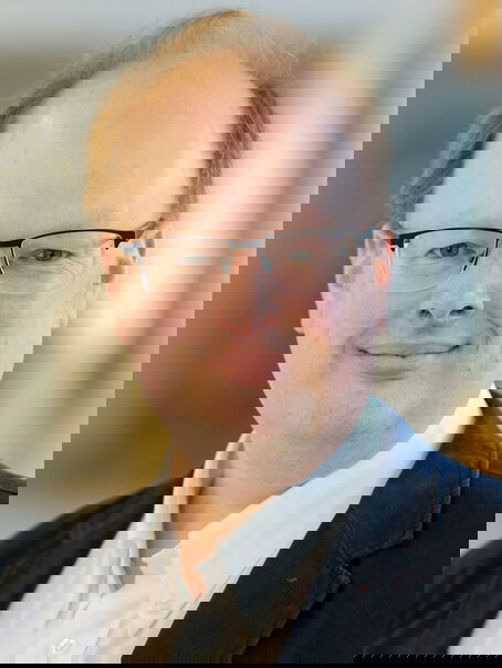 Das Bild zeigt eine männlich lesbare Person mit kurzen mittelblonden Haaren und einer tiefen Stirn, einer Brille mit dunkler, halber Fassung, weißem Hemd und schwarzem Sakko.