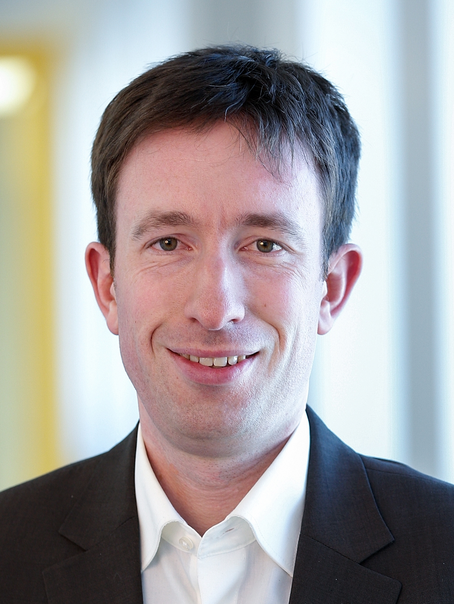 Das Bild zeigt eine männlich lesbare Person ohne Bart mit dunklen kurzen Haaren, weißem Hemd und dunklem Sakko.