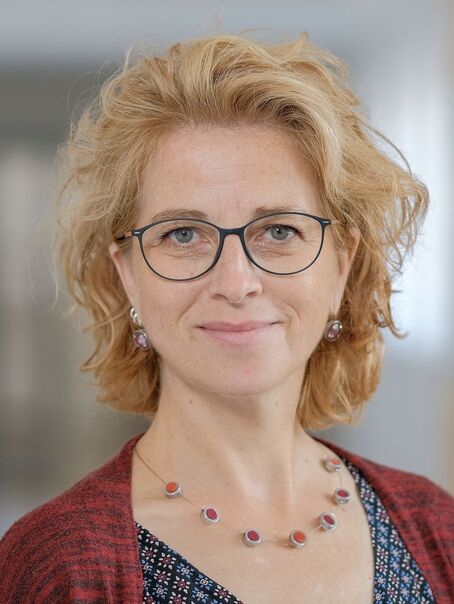 Das Bild zeigt eine weiblich lesbare Person mit mittellangen blonden Haaren, einer Brille mit dunkler Fassung, einer roten Strickjacke und einer blauen Bluse.