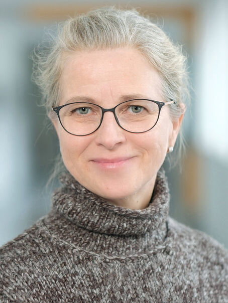 Das Bild zeigt eine weiblich lesbare Person mit zurückgebundenen blondgrauen Haaren und einer Brille mit schwarzer Fassung.