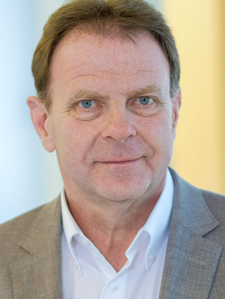 Das Bild zeigt eine männlich lesbare Person mit kurzen, mittelbraunen Haaren im Portrait. Die Person trägt keinen Bart, ein weißes Hemd und ein hellbraunes Sakko. 