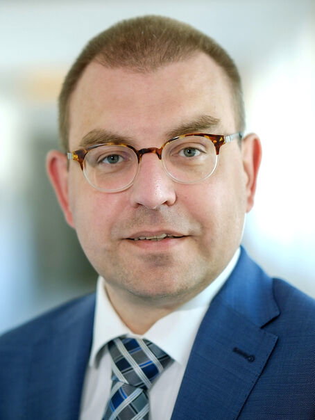 Eine männlich gelesene Person mit kurzen Haaren und einer Brille, mit einem blauen Sakko und gestreifter Krawatte.