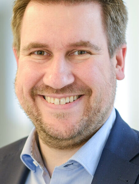Das Bild zeigt eine männlich lesbare Person mit kurzen, mittelbraunen Haaren im Portrait. Die Person trägt einen Bart, ein hellblaues Hemd und ein dunkelblaues Sakko.
