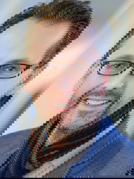 Das Bild zeigt eine männlich lesbare Person mit schwarzem Vollbart, dunklen kurzen Haaren und einer Brille mit schwarzer Fassung. Die Person trägt einen dunklen Schal, ein hellblaues Hemd und ein blaues Sakko. 