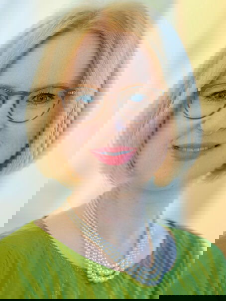 Das Bild zeigt eine weiblich lesbare Person mit blondem Pagenschnitt, brauner Brille, einer Perlenkette und einem grünen Oberteil.
