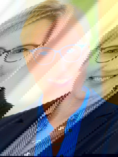 Das Bild zeigt eine weiblich lesbare Person mit hellblonden kurzen Haaren, einer Brille mit dunkler Fassung, einer blauen Bluse und einem dunklen Sakko. 