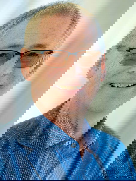 Das Bild zeigt eine männlich lesbare Person ohne Bart mit kurzen graumelierten Haaren, einer leicht getönten Brille, blauem Hemd und blauem Sakko.