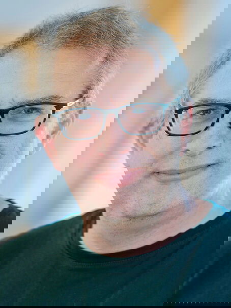 Eine männlich lesbare Person ohne Bart mit kurzen, grauen Haaren, einer Brille mit schwarzer Fassung und einem schwarzen Pullover.