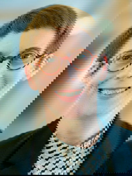 Eine weiblich gelesene Person mit dunklen kurzen Haaren, einer Brille mit dunkler Fassung und einem dunklen Jackett.