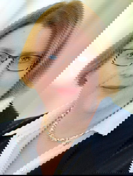 Das Bild zeigt eine weiblich lesbare Person mit mittelbraunem Pagenschnitt, einer Brille mit dunklem Rahmen, einer weißen Bluse und einem dunklen Jackett.