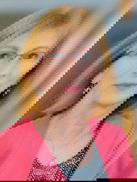 Das Bild zeigt das Portrait einer weiblich lesbaren Person mit schulterlangen rötlichen Haaren, einer Brille und einem hellroten Jackett.