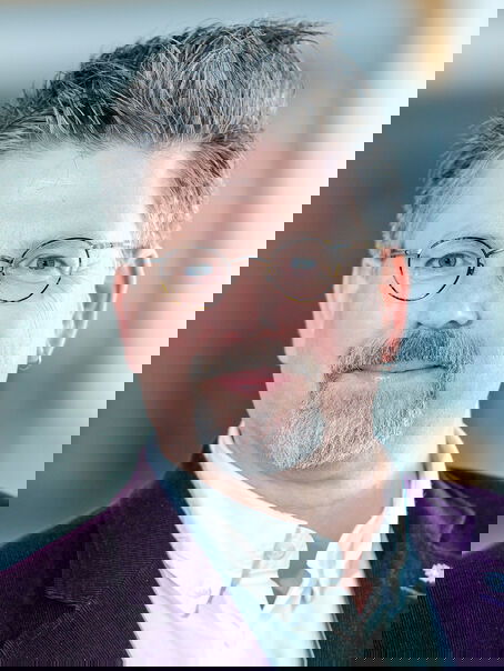 Das Bild zeigt eine männlich lesbare Person mit grauem Bart und schwarz-grauen Haaren und einer Brille mit dunkler Fassung. Sie trägt ein weißes Hemd und ein dunkles Sakko.