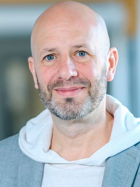 Das Bild zeigt eine männlich lesbare Person mit grauem Bart und fast kahlem Kopf, einem weißen Kapuzen-Pullover und einem hellblauen Sakko.