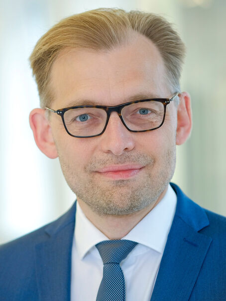 Das Bild zeigt eine männlich lesbare Person ohne Bart mit kurzen dunkelblonden Haaren, einer Brille mit dunkler Fassung, weißem Hemd, blauer Krawatte und blauem Sakko. 
