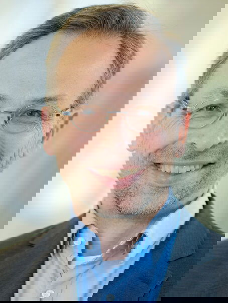 Das Bild zeigt eine männlich lesbare Person mit Drei-Tage-Bart, dunklenen graumelierten Haaren, einer Brille, hellblauem Hemd und dunklem Sakko.