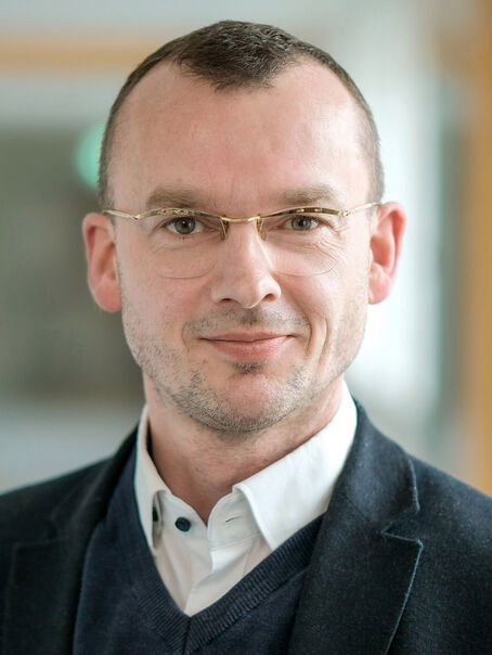 Eine männlich gelesene Person mit sehr kurzen dunklen Haaren, einer Brille ohne Fassung, einem dunklen Pullover und weißem Hemd.