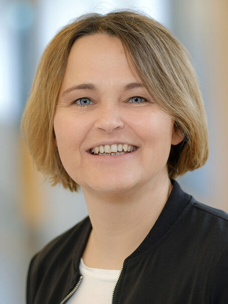 Eine weiblich gelesene Person mit kinnlangem dunkelblonden Haar, weißem Oberteil und schwarzer Jacke.