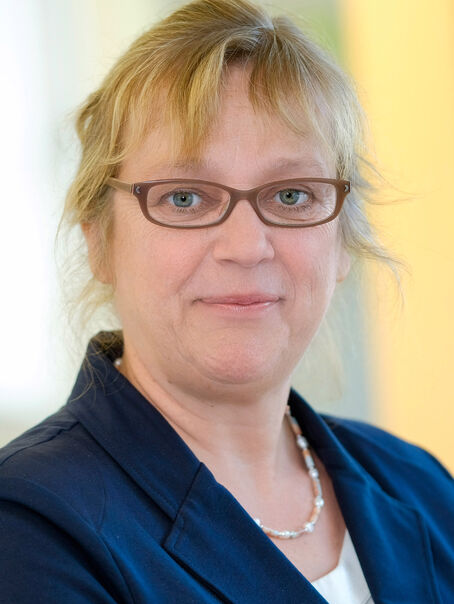 Das Bild zeigt eine weiblich lesbare Person mit längeren, hochgesteckten blonden Haaren, einer Brille mit dunkler Fassung, einem weißen Oberteil und einem dunkelblauen Sakko.