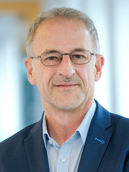 Eine männlich gelesene Person mit kurzen grauen Haaren, Brille und kurzem grauen Bart. Er trägt ein dunkelblaues Sakko und ein hellblaues Hemd. 