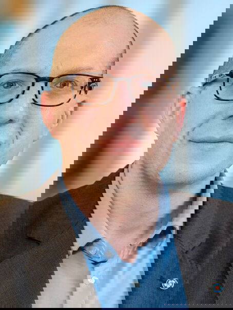 Das Bild zeigt eine männlich lesbare Person mit kurzen dunklen Haaren, einer dunklen Brille, hellblauem Hemd und dunklem Sakko.