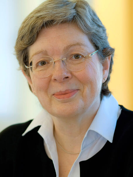 Das Bild zeigt eine weiblich lesbare person mit kurzen, graumelierten Haaren, einer Brille ohne Fassung, einer weißen Bluse und einem dunklen Jackett.
