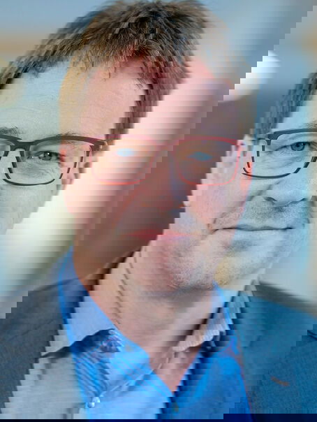 Eine männlich gelesene Person mit kurzen mittelbraunen Haaren, einer mittelbraunen Brillenfassung, blauem Hemd und blauem Sakko.