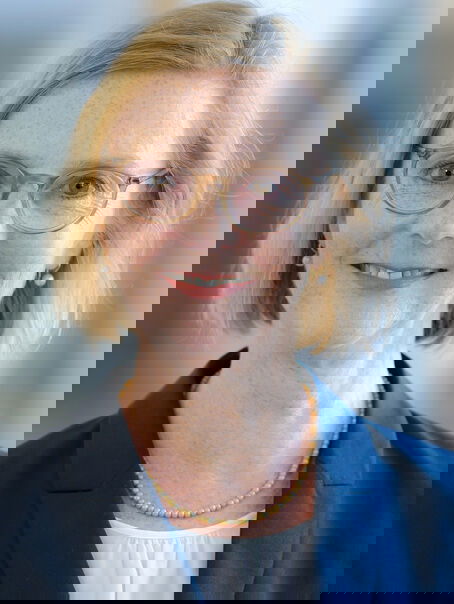 Weiblich lesbare Person mit kinnlangen blonden Haaren, Brille, weißem Oberteil und blauem Jackett.