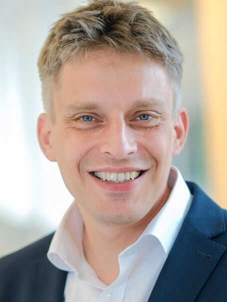 Das Bild zeigt eine männlich lesbare Person ohne Bart mit kurzen graumelierten Haaren, weißem Hemd und dunklem Sakko.
