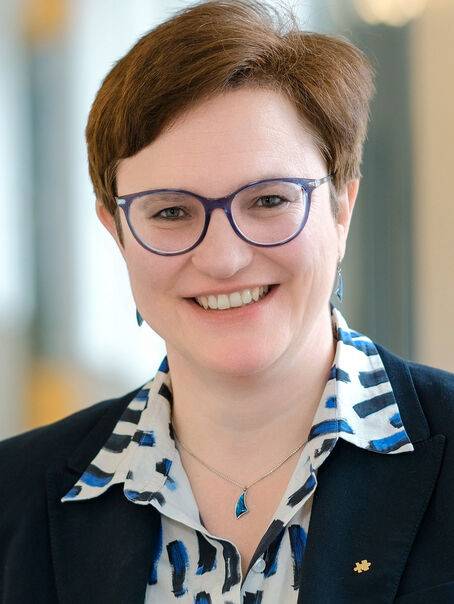 Eine weiblich gelesene Person mit kurzen braunen Haaren und Brille, in gemusterter Bluse und dunklem Sakko.