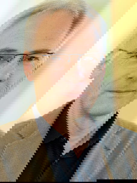 Das Bild zeigt eine männlich lesbare Person ohne Bart mit kurzen grauen Haaren, einer Brille ohne Fassung, einem schwarzen Hemd und einem mittelbraunen Sakko.