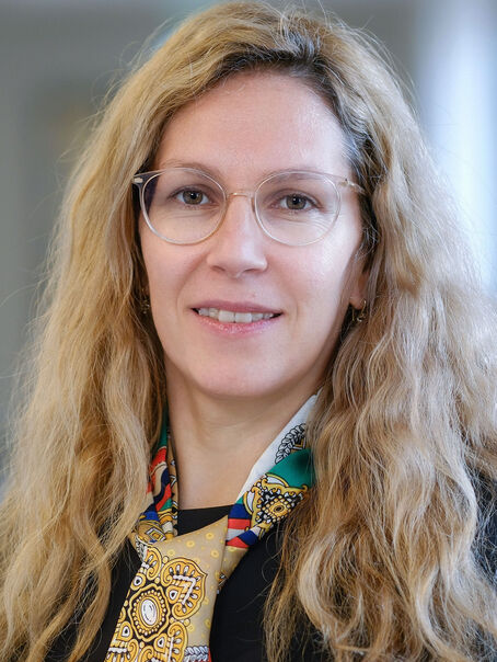 Das Bild zeigt eine weiblich lesbare Person mit langem blonden Haar, einer Brille ohne Fassung, einem gemusterten Schal und einem dunklen Oberteil. 