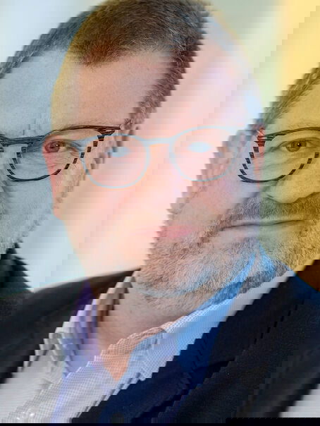 Das Bild zeigt eine männlich lesbare Person mit kurzem graumelierten Bart, kurzen dunklen Haaren, einer Brille mit dunkler Fassung, einem hellblauen Hemd und dunklem Jackett.
