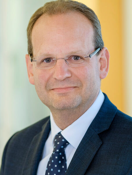 Das Bild zeigt eine männlich lesbare Person ohne Bart mit kurzen dunklen Haaren, einer Brille ohne Fassung, einem weißen Hemd, einem dunklen Sakko und einer blauen Krawatte mit weißen Punkten.