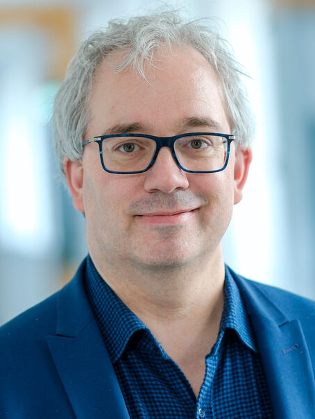 Eine männlich gelesene Person mit kurzem grauen Haar, einer dunklen Brille, blauem Hemd und blauem Sakko. 