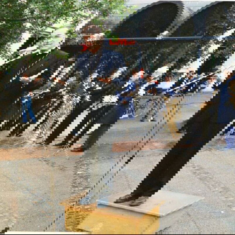 Das Bild zeigt eine männlich lesbare Person in schwarzem Anzug, die auf einem Platz auf einem kleinen Podest stehend in ein Mikrophon spricht.