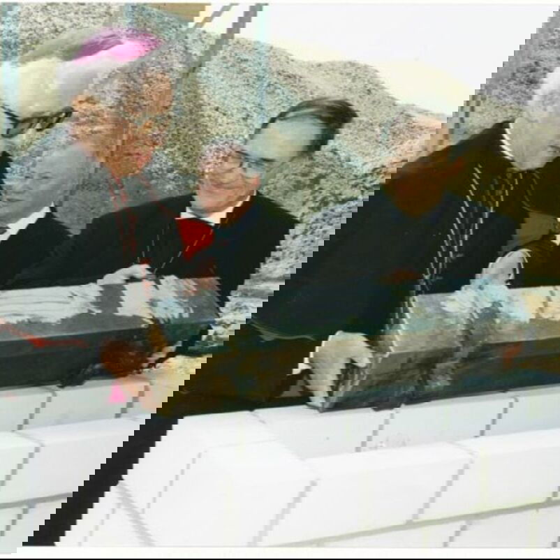 Das Bild zeigt zwei männlich lesbare Personen, eine in schwarzem Anzug, die andere im Talar, die gemeinsam einen Grundstein setzen.