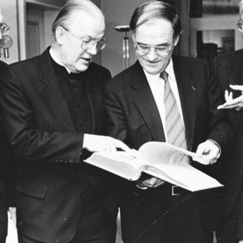 Das Bild zeigt zwei männlich lesbare Personen, die nebeneinander stehen und gemeinsam in ein großes aufgeschlagenes Buch schauen, das sie gemeinsam festhalten.