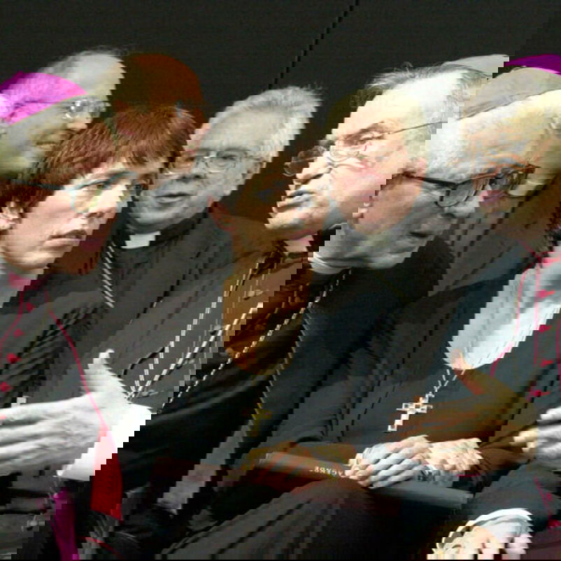 Das Bild zeigt eine weiblich lesbare Person mit kurzen dunklen Haaren dicht bedrängt von einer Gruppe katholischer Geistlicher, die um sie herum sitzen und mit ihr sprechen.