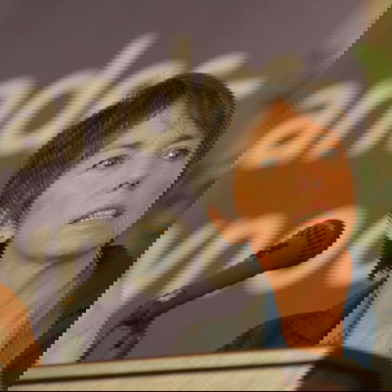 Das Bild zeigt eine weiblich lesbare Person mit kurzen dunklen Haaren, die in ein Mikrophon spricht.