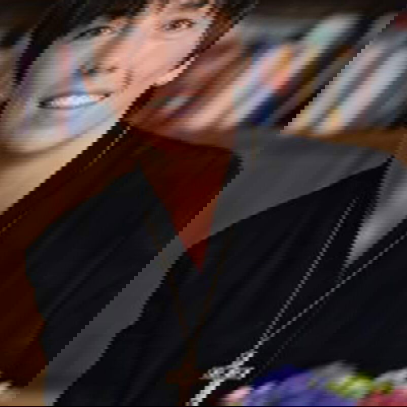 Das Bild zeigt eine weiblich lesbare Person mit dunklen kurzen Haaren, dunklem Oberteil, sitzend vor einer Bücherwand. Im Vordergrund steht ein Blumenstrauß.