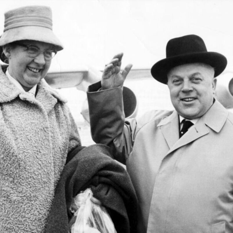 Das Schwarzweiß-Bild zeigt je eine männlich und eine weiblich lesbare Person mit Hut und Mantel vor einem Flugzeug.