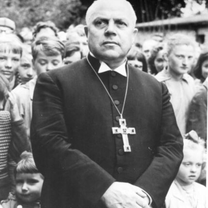 Das Schwarzweiß-Bild zeigt eine männlich lesbare Person in Talar und einer Kette mit einem großen Kreuz vor der Brust.