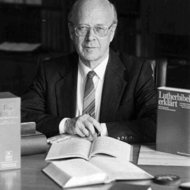 Das Schwarzweiß-Bild zeigt eine männlich lesbare Person mit kurzen hellen Haaren, einer Brille, weißem Hemd, Krawatte und dunklem Sakko, sitzend mit aufgeschlagenen Büchern an seinem Schreibtisch.